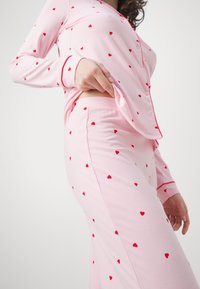 Ljusrosa pyjamasuppsättning med små röda hjärtan, långärmad och med knappslå, samt matchande åtsittande byxor. Mjuk textur.