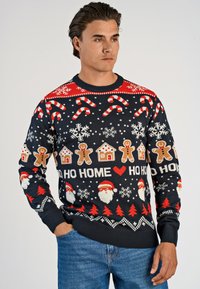 Man som bär en marinblå jultröja med godiskäppar, snöflingor, pepparkaksgubbar, tomtens ansikten, röda träd och texten "HO HO HOME".