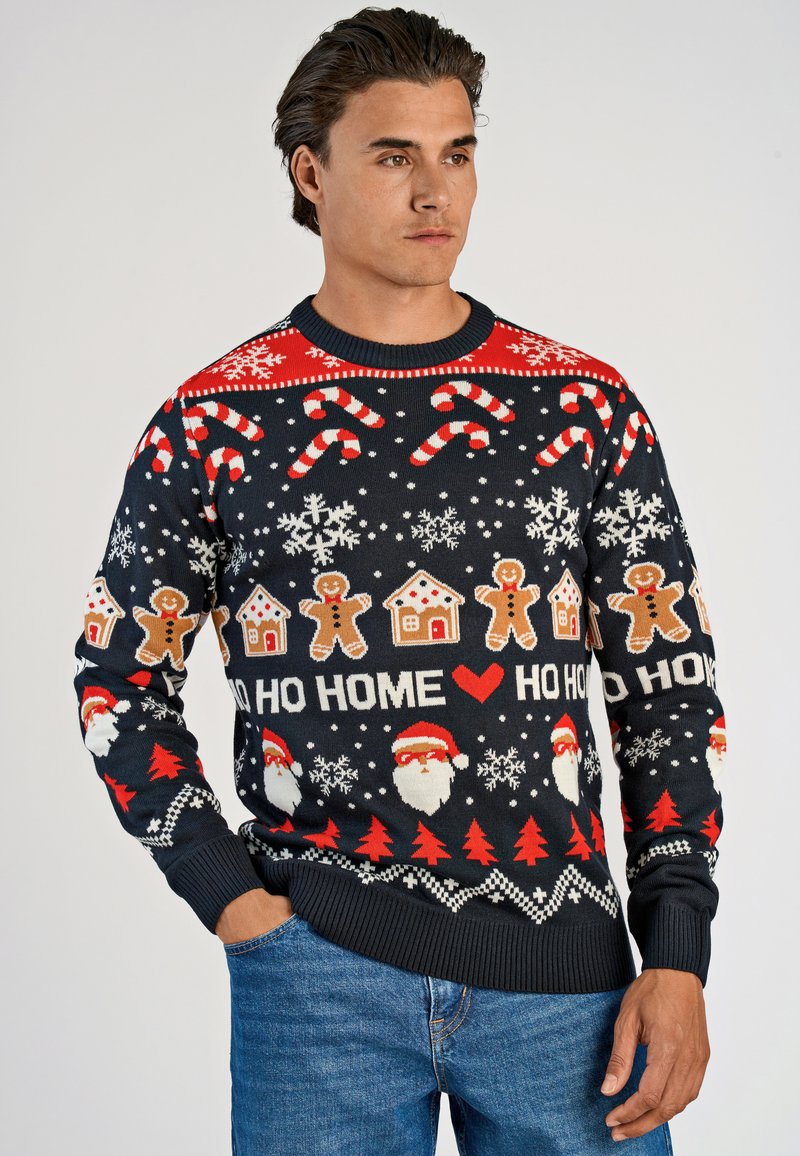 Man som bär en marinblå jultröja med godiskäppar, snöflingor, pepparkaksgubbar, tomtens ansikten, röda träd och texten "HO HO HOME".