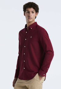 Bordeaux-kleurige overhemden met lange mouwen, voorzien van een subtiele logo op de borst en een gebogen zoom. Gdracht met beige broek.