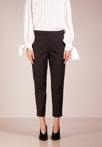 Femme portant un pantalon noir court, des talons noirs, et une blouse blanche à manches longues avec des rayures roses et des poignets noués, se tenant sur un fond neutre.