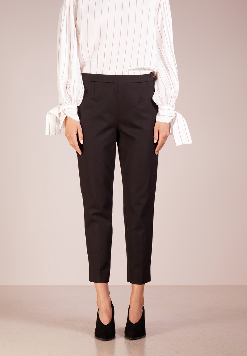 Femme portant un pantalon noir court, des talons noirs, et une blouse blanche à manches longues avec des rayures roses et des poignets noués, se tenant sur un fond neutre.