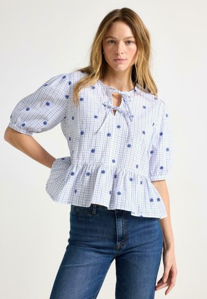 Ung kvinde med blondt hår iført en hvid bluse med blåt blomsterternet mønster, pufærmer, bindesnøre ved halsen og blå jeans, stående mod en hvid baggrund.