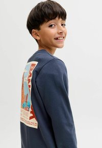 Ragazzo con capelli corti e scuri che indossa una felpa blu scuro con una grafica colorata "Bangkok" sulla schiena, sorridente e che guarda oltre la spalla.