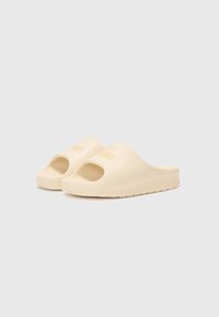 Lys beige plast sandaler med åpen tå og sideutskjæringer, med en subtil preget logo på stroppen og en teksturert såle.