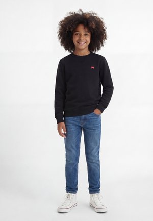 Παιδί με σγουρά μαλλιά που φοράει μαύρο φούτερ, μπλε τζιν και λευκά αθλητικά παπούτσια, στέκεται με το χέρι στην τσέπη και χαμογελάει.