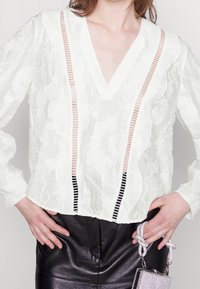 Blusa bianca testurizzata con scollo a V profondo, caratterizzata da bordi arricciati e strisce verticali a contrasto. Abbinata a pantaloni in pelle nera.