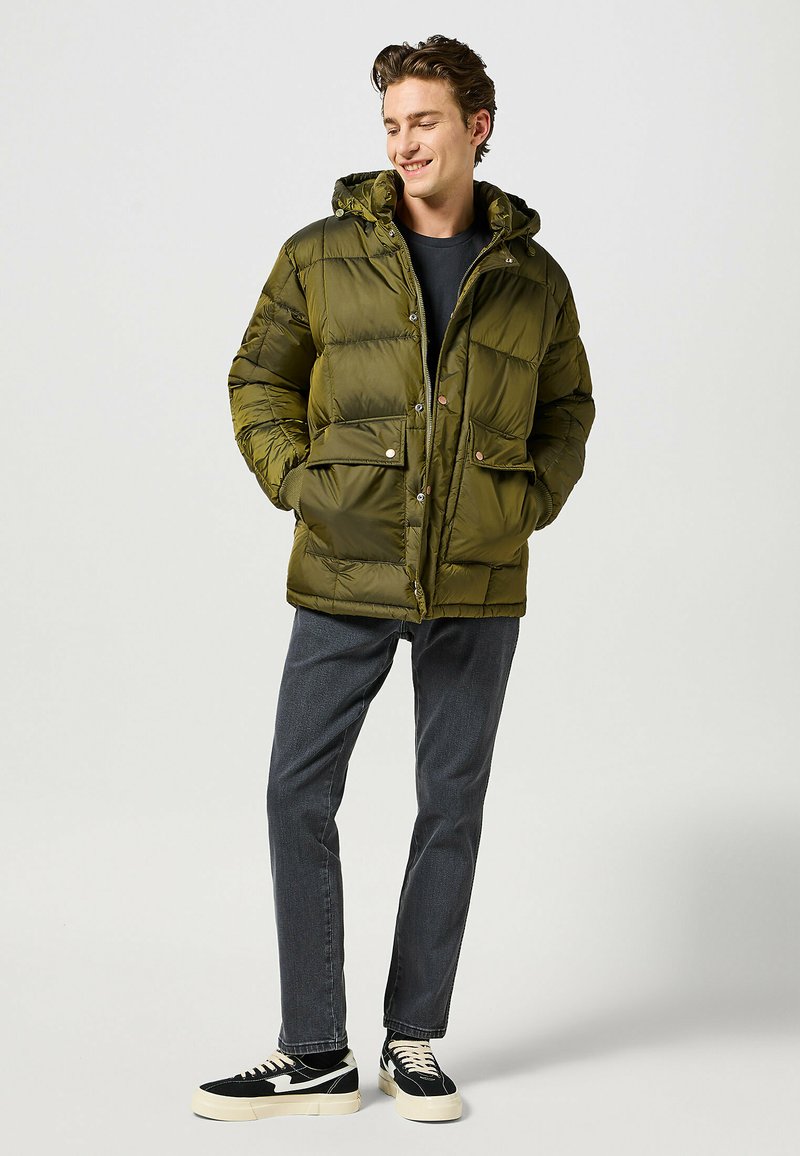 Olíva zöld puffos dzseki kapucnival, elöl zipzárral, két elülső zsebbel és rugalmas mandzsettákkal, sötét ing és szürke farmer felett viselve.