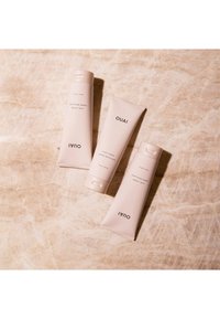 Trois tubes cosmétiques rose clair étiquetés "OUAI" sur une surface beige texturée. Deux tubes sont verticaux ; un est légèrement incliné.