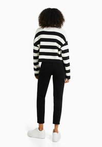 Suéter cortado de rayas blancas y negras con textura acanalada, combinado con jeans negros y zapatillas blancas. La vista trasera muestra un ajuste relajado.
