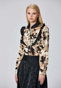 Blouse à motif floral en beige, noir et crème avec des accents volantés et une taille sombre. Manches longues, texture lisse et design avec col.
