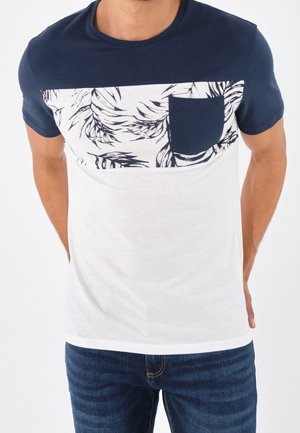 Homme portant un t-shirt bleu marine et blanc à manches courtes avec un motif de feuilles noires sur la poitrine et une poche bleu marine, associé à un jean bleu.