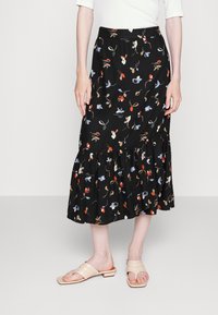 Saia preta floral com padrões coloridos, bainha com babados e cintura ajustada; combinada com sandálias de dedo em bege.