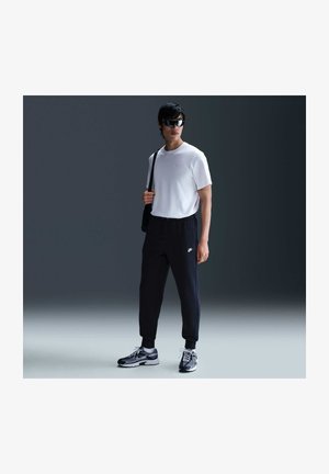 Homme portant des lunettes de soleil noires, un t-shirt blanc, un pantalon de survêtement noir Nike, des baskets noires et un sac noir en bandoulière, debout à l’intérieur.