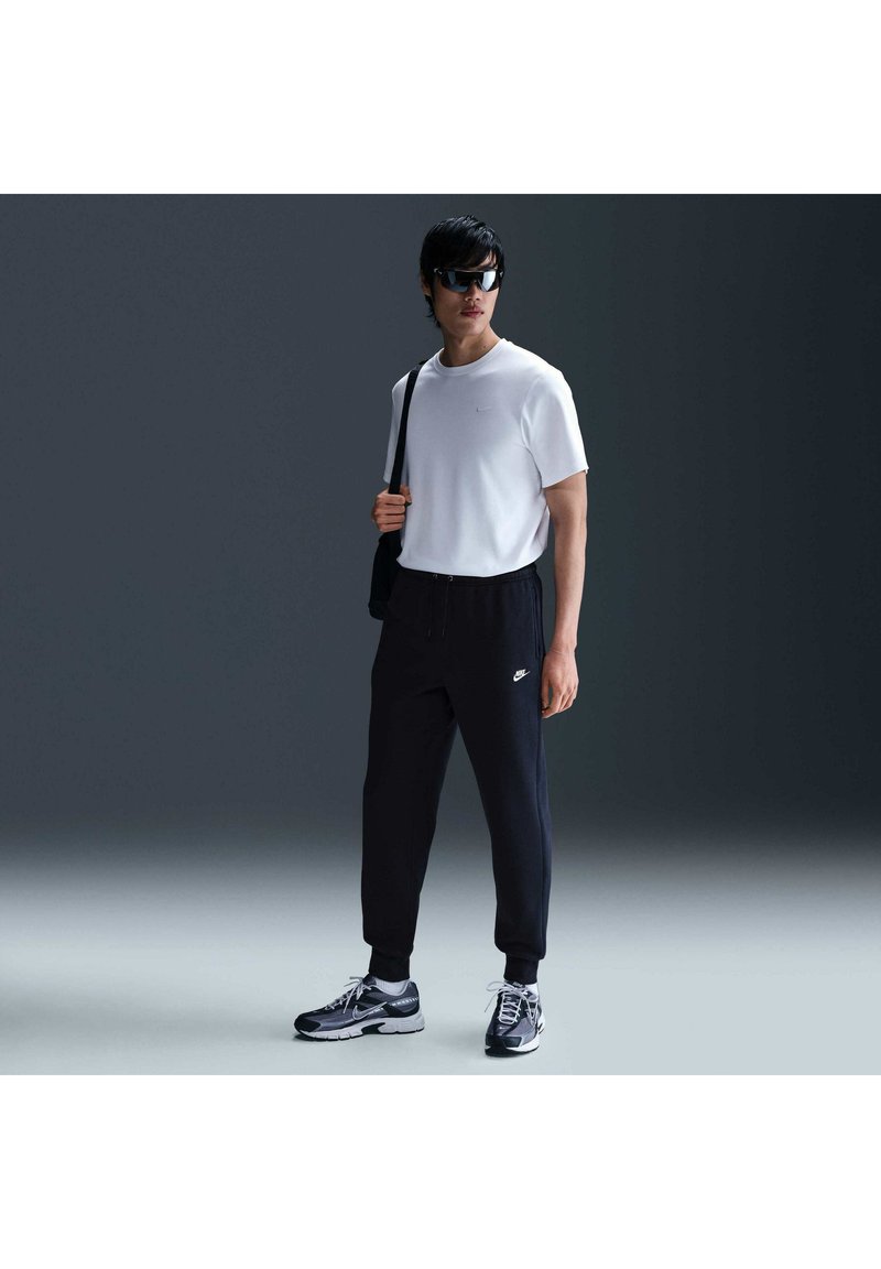 Uomo con occhiali da sole neri, t-shirt bianca, pantaloni da tuta neri Nike, sneakers nere e borsa a tracolla nera, in piedi all'interno.