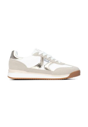 Zapatilla blanca y beige con detalles metálicos plateados, diseño con cordones y suela de goma marrón, mostrada de lado sobre un fondo blanco.