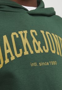 Sudadera verde con texto amarillo "JACK & JONES intl. desde 1990" llevada sobre una camisa blanca, mostrando un primer plano del área del pecho.