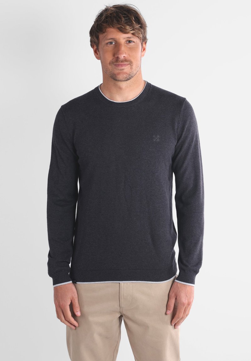 Homme portant un pull à manches longues gris foncé et un pantalon beige, se tenant devant un fond clair uni, tourné vers l'avant avec une expression neutre.