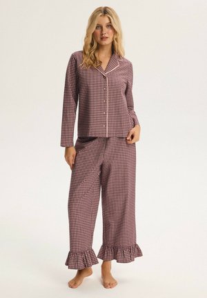 Blonde woman wearing burgundy and white checkered pajama set with buttoned long-sleeve top and ruffled wide-leg pants, standing barefoot.
