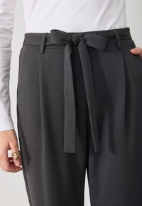 Detailní záběr na tmavě šedé vysoké kalhoty s látkovým páskem uvázaným do mašle, nošené s bílým topem s dlouhým rukávem.