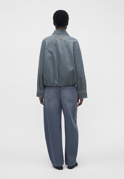 Personne debout de dos, portant une veste en jean ample et un pantalon en jean à jambes larges sur un fond clair uni.