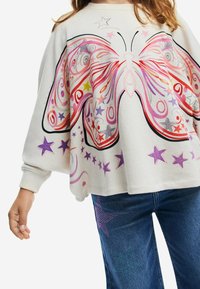 Felpa grigia chiaro con una grande grafica di farfalla in spirali rosa e rosse; abbinata a jeans blu con decorazioni a forma di stella in turchese e viola.