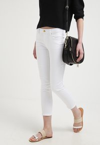 Mujer vestida con pantalones cortos ajustados blancos, suéter corto negro, sandalias de slide beige y plateadas, llevando un bolso de cuero negro cruzado.