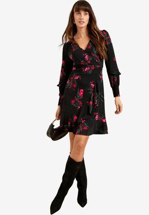 Schwarzes Wickelkleid mit langen Ärmeln, das mit pinkfarbenen Blumenmustern und Rüschendetails versehen ist, kombiniert mit knielangen schwarzen Stiefeln und einer kleinen Handtasche.