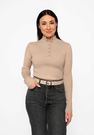 Rippstrick-beige Langarmtop mit hohem Ausschnitt und Knopfdetails, kombiniert mit hoch taillierten schwarzen Denim-Jeans und einem hellen Gürtel.