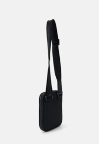 Černá crossbody taška s hladkým povrchem, vybavená zipovým uzávěrem a popruhem s vyraženým logem. Lehký design s nastavitelnou délkou.