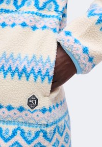 Main dans la poche d'une veste polaire crème avec des motifs géométriques bleus et roses et un petit patch logo Kari Traa noir et blanc.
