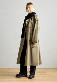 Palton lung bej cu nasturi dubli, asortat cu o eșarfă neagră. Un model poartă pantaloni negri și cizme negre masive.