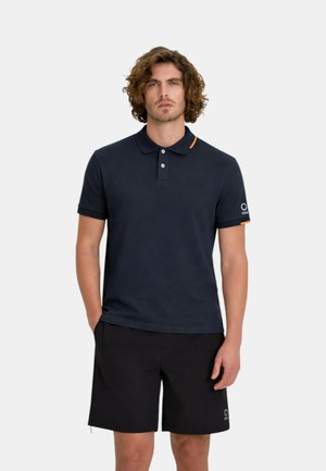 Uomo con i capelli ricci che indossa una polo blu navy con dettagli arancioni e pantaloncini neri, in piedi con una mano in tasca.