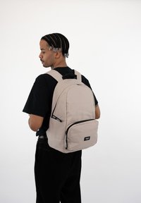 Krém színű hátizsák állítható pántokkal, elülső cipzáras zsebbel és téglalap alakú formával, egy személy hátán viselve fehér háttér előtt.