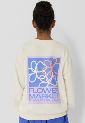 Person mit zu einem Dutt gebundenem Haar, trägt ein cremefarbenes Sweatshirt mit einem blauen und rosa Blumenmuster und dem Text "FLOWER MARKET PLANT GROW BLOOM".