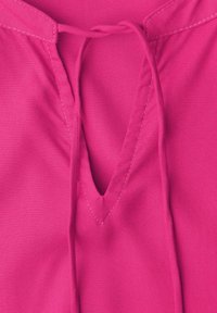 Tissu rose vif avec une ouverture en V et des cordons noués, présentant des surpiqûres visibles le long des bords.