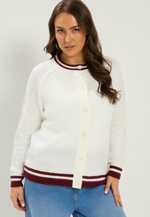 Kvinde med langt mørkt hår iført en hvid strikket cardigan med bordeaux og hvide stribede manchetter og kant, kombineret med blå jeans.