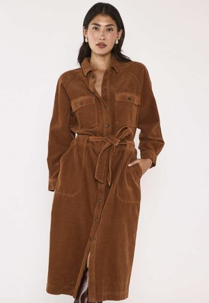 Donna che indossa un lungo vestito marrone in velluto a coste con bottoni frontali, due tasche sul petto, tasche laterali e cintura annodata in vita.