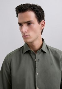 Camisa verde oliva de botones con cuello, que presenta un pequeño logo en el lado izquierdo del pecho, hecha de una tela suave y con un corte relajado.
