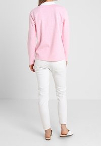 Helles rosa Sweatshirt mit Rundhalsausschnitt, weicher Textur, kombiniert mit weißen eng anliegenden Jeans und Slippern. Rückansicht dargestellt.