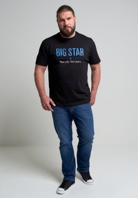 Camiseta de algodón negra con texto azul "BIG STAR", combinada con jeans de mezclilla azul y zapatillas negras con suelas blancas. Diseño simple y casual.