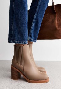 Skórzane brązowe botki na blokowym obcasie noszone z niebieskimi dżinsami, stojące na jasnoszarym podłożu, w tle widoczna brązowa zamszowa torba.