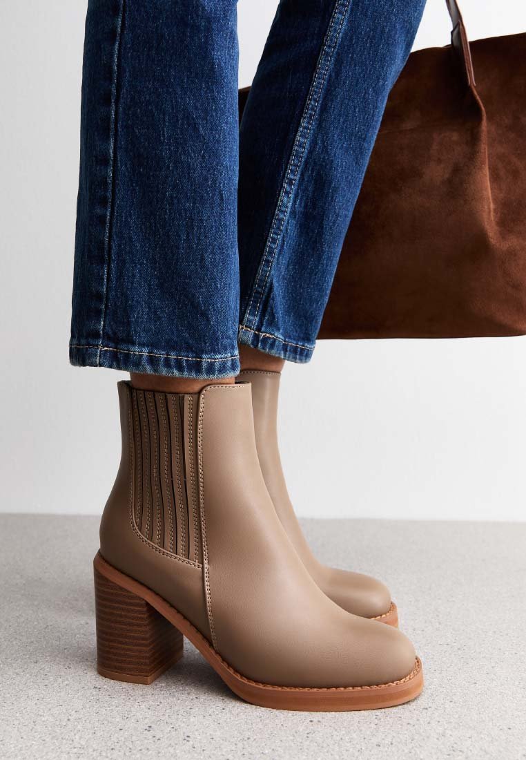Skórzane brązowe botki na blokowym obcasie noszone z niebieskimi dżinsami, stojące na jasnoszarym podłożu, w tle widoczna brązowa zamszowa torba.