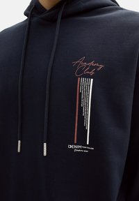 Granatowa bluza z kapturem ze sznurkiem, z wyraźnym graficznym nadrukiem "Academy Club" w kolorach burgundowym, białym i czarnym na piersi. Miękka struktura materiału.