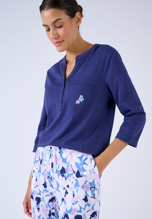 Top de manga larga color azul marino con escote henley y bolsillo frontal, con bordado floral. Combinado con pantalones de patrón floral en azul claro.