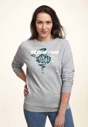 Grijze crewneck sweatshirt met witte en teal grafische tekst en ontwerpen. Het model draagt het met blauwe jeans. Zachte textuur en een relaxte pasvorm.