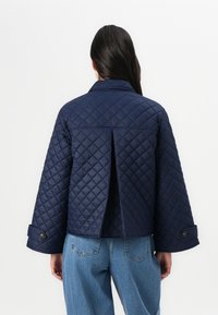 En person står med ryggen til, iført en marineblå quiltet jakke med splitt bak, sammen med lyseblå jeans, mot en enkel hvit bakgrunn.