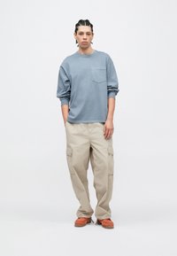 Μπλε πουκάμισο με μακ ριά μανίκια και τσέπη στο στήθος, σε συνδυασμό με μπεζ cargo παντελόνια και πορτοκαλί παπούτσια. Το σύνολο έχει χαλαρή εφαρμογή και απαλή υφή.