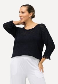 Marinblått stickad tröja med en lös passform, som har en texturerad design, trekvartslånga ärmar och fransdetaljer i nederkant. Kombinerad med vita byxor.