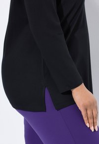 Zijaanzicht van een persoon in een zwarte lange mouw top met een zijsplit en paarse leggings, hand rustend op de dij.